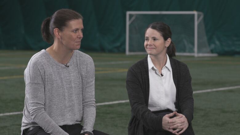 Two women sit on chairs on an empty soccer field. 