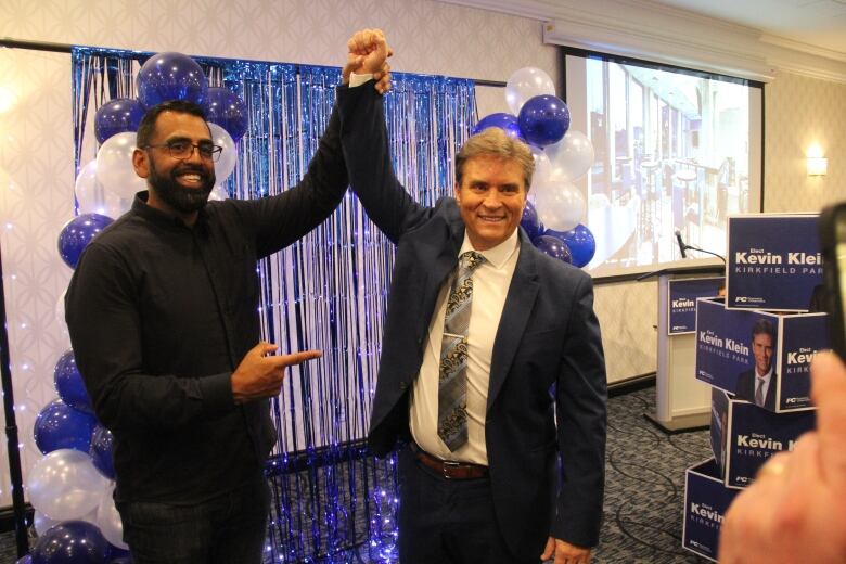 Two men holding each other's arm up with balloons in the background.