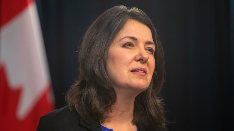 A woman wearing a black blazer is speaking in front of a Canadian flag.