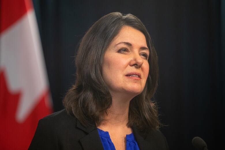 A woman wearing a black blazer is speaking in front of a Canadian flag.