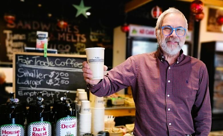 Man with glasses holds up a paper coffee cup inside a cafe.