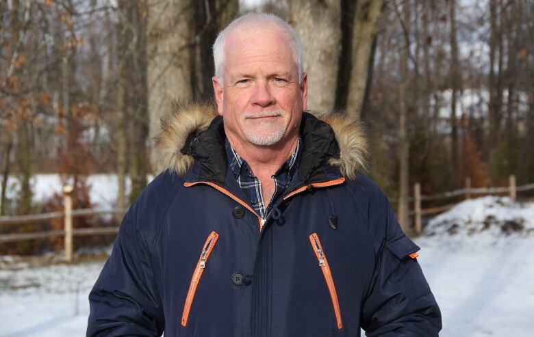 A balding man white white hair and a goatee stands in his backyard. He has a slight smile and is wearing a dark blue winter jacket with orange zippers and fur-lined hood. There is snow on the ground and trees behind him. 