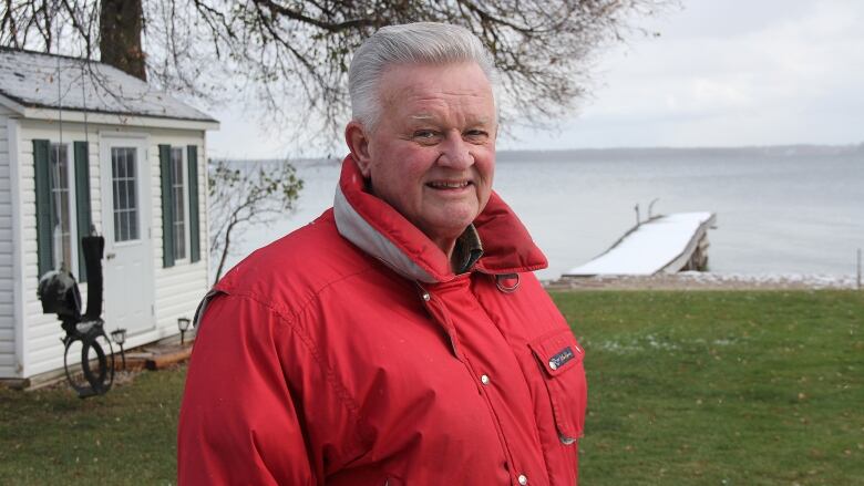 A man with white hair stands in his backyard, facing the camera. Behind him a wooden dock stretches out into the water. There is some snow on the ground.