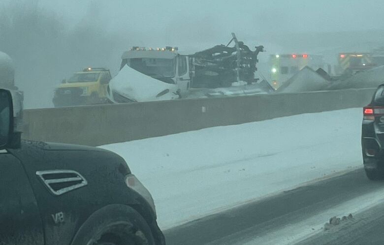 Cars drive past wrecked vehicles seen behind a highway median in a snowstorm.