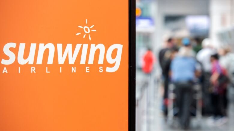 Passengers walk near an orange airline sign that reads, 'Sunwing Airlines.' 