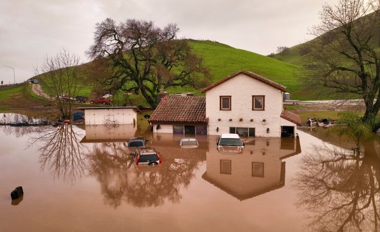 Submerged vehicles and a house in muddy water are shown in front of a house located below rolling green hills.