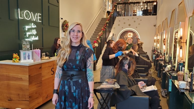 Dana Lyseng stands in middle of her busy hair salon.  She has long blond hair and clear framed glasses. She is wearing a navy blue dress with a light blue floral print.  The dress has short sleeves which reveal her heavily tattooed arms.    