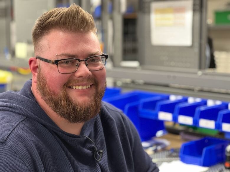 Jace Staples smiles while sitting at his work station.   He has straight white teeth,  reddish short hair with a spikey cut and a thick neatly trimmed heard.  He was dark framed glasses and has a piercing in his eyebrow.     