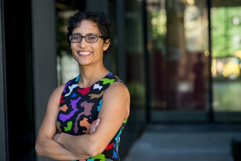 Dr Monika Dutt stands with her arms crossed outside a modern building, smiling into the camera.  She has short back hair and  dark framed glasses.  She's wearing a black dress with a random pattern of red, white, green and blue shapes that have jagged edges. She has light brown skin.  