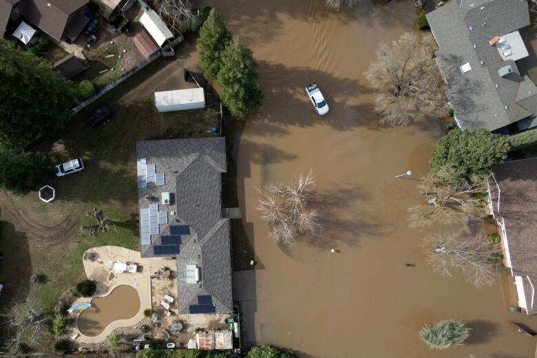 A drone view above a flooded street shows a car driving through a brown mini lake and a muddy brown swimming pool.