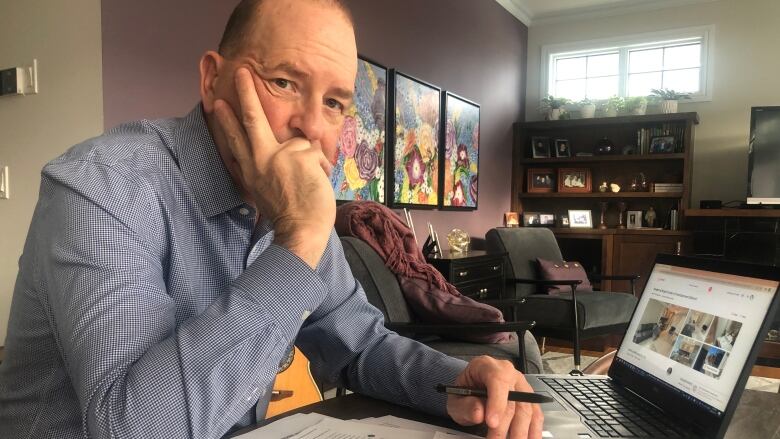 A balding man with a blue button up shirt sits at a table staring at a laptop screen. There are lots of papers scattered across the table. 