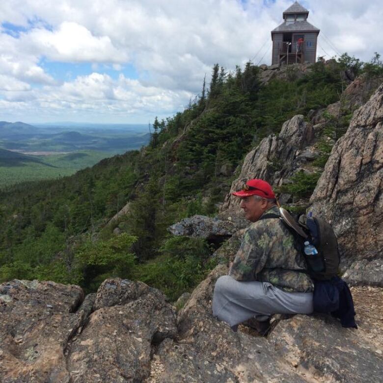 An Indigenous man in a red hat sits on a mountain side. 