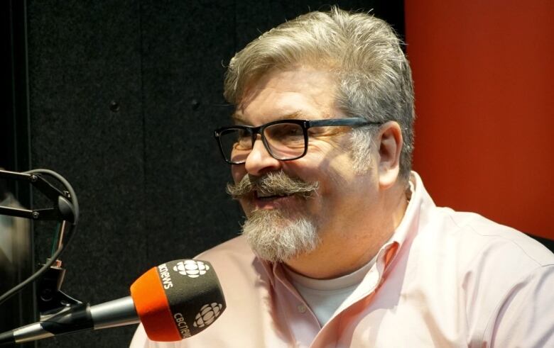 A man wearing glasses and a pink shirt sits inside a CBC Kitchener-Waterloo radio booth.