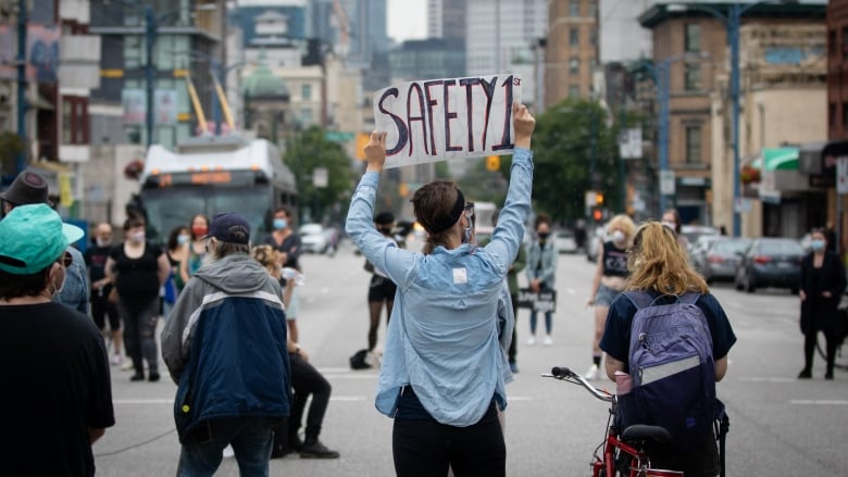 A group of four people stand in the middle of a Vancouver street, with their backs to the camera. One is holding a sign reading 'Safety 1st' above their head.