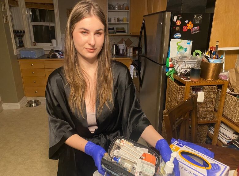 A woman stands in her kitchen in a black robe holding a prescription opioid kit, including sanitizing wipes, bottles and needles.