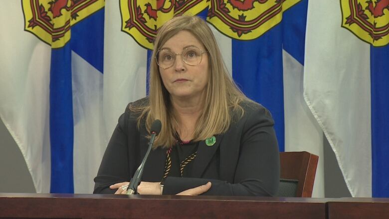 A woman sits at a table and microphone in front of a row of Nova Scotia flags.