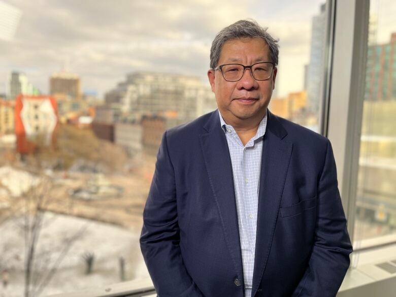 A man in a navy blazer stands in front of a 5th floor office window with the skyline of Toronto's east downtown behind him. 