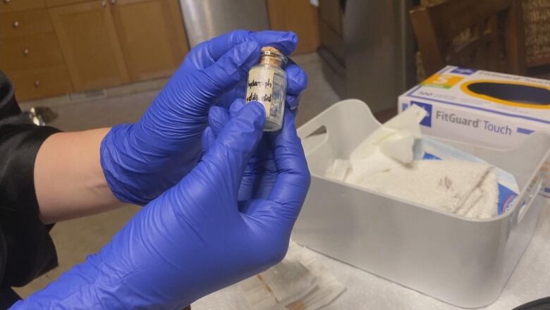 Latex-gloved hands hold a small, clear vial labelled as hydromorphone. Other medical supplies sit in the background.