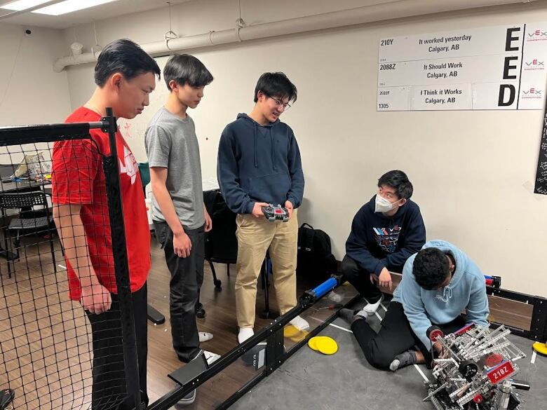 five boys stand in a semi circle around a robot with wheels. they are talking 