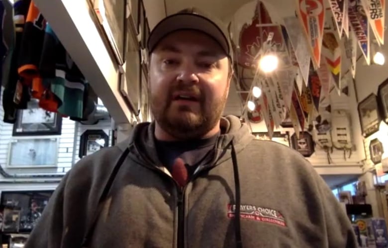 A man in jacket and wearing a ball cap speaks inside a shop full of merchandise.