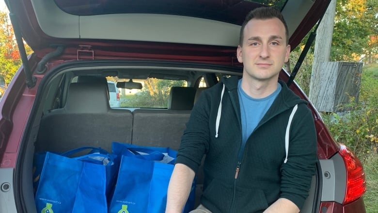 A white man with short brown hair wearing light coloured jeans, a blue t-shirt, and a dark hoodie sits on the edge of his compact car's trunk, with the hatch open. He is sitting beside two blue cloth shopping bags.