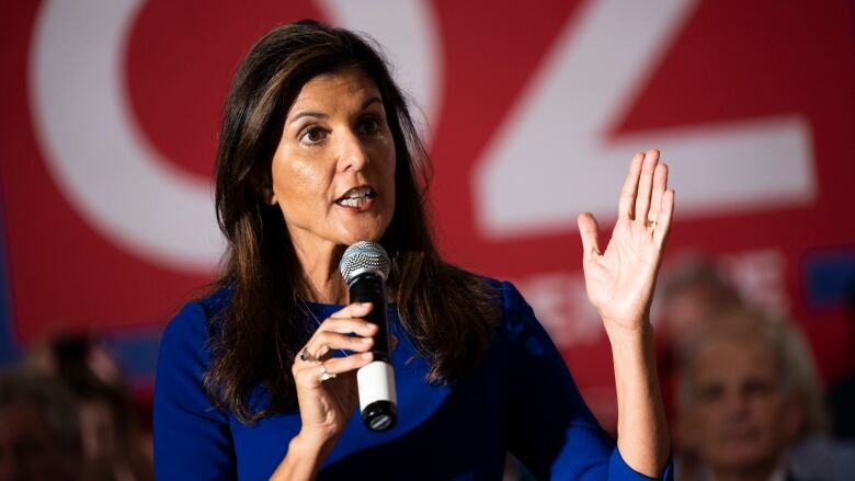 A woman is shown standing and speaking into a microphone.