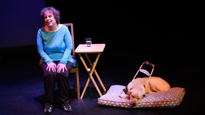 A woman wearing a blue shirt sits in a chair beside a creamy white labrador retriever on a darkly lit stage.