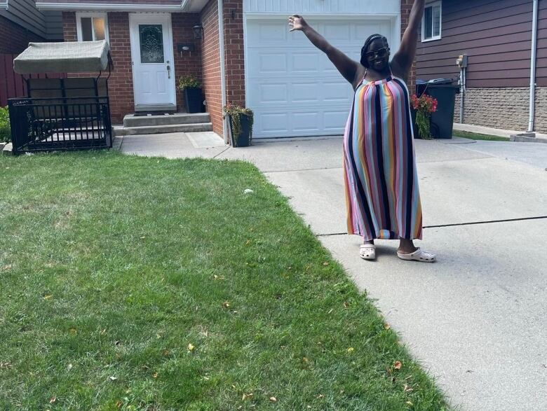 A woman stands in front of a house with her arms raised in excitement.