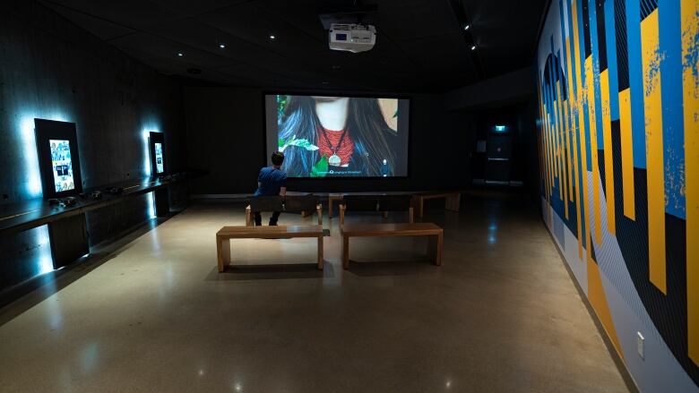 A man watches a video in a darkened room. A screen plays in front of the man. 