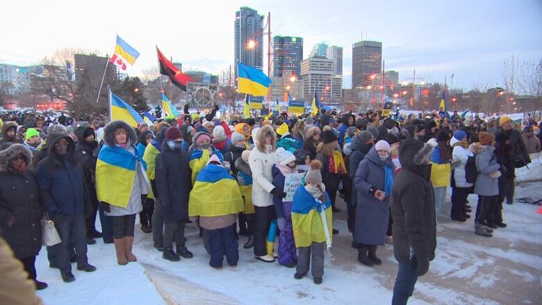 Hundreds of people carrying Ukrainian flags stand together, with downtown Winnipeg in the background.