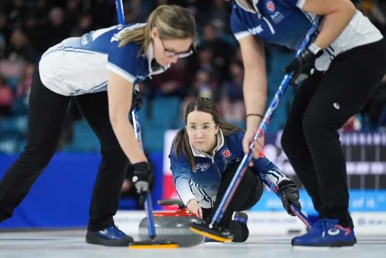 A female curler slides the rock along the ice as two other curlers sweep.