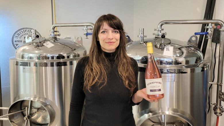 A woman holding a bottle of pink kombucha in her left hand is shown standing in front of two steel brewing vats. 