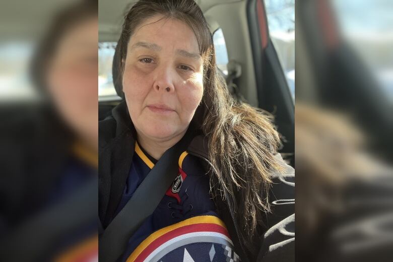 A woman with long dark hair looks at the camera while sitting in a car.