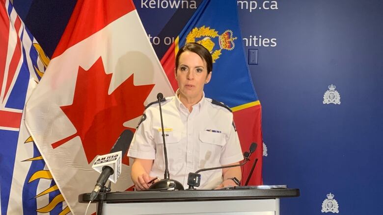 A woman in police uniform speaks on the podium, with flags and RCMP sign on the background.