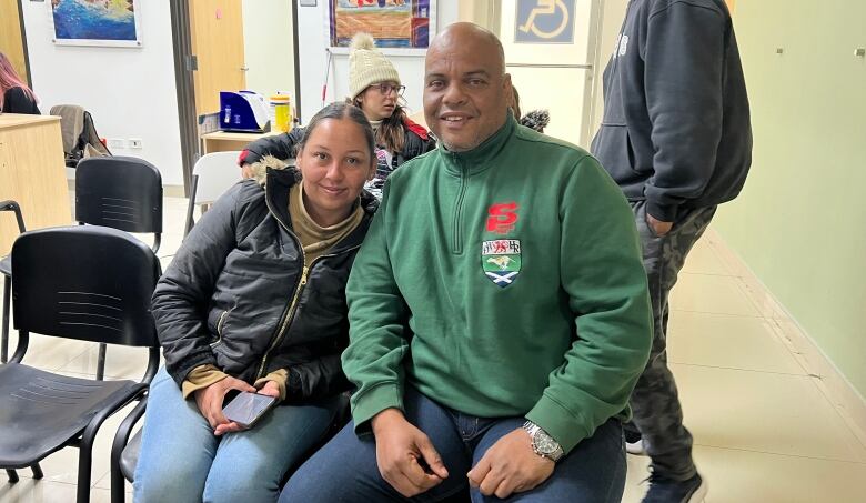 A woman in a black jacket and a man in a green sweatshirt huddle together to pose for a portrait in the lobby of an administrative building.