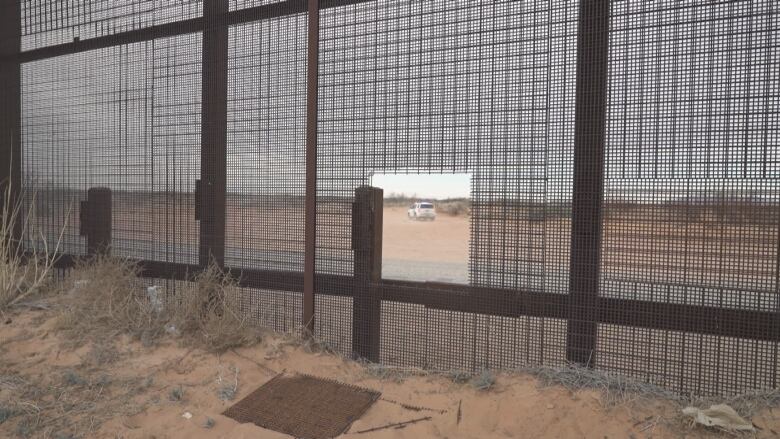 A square hole is cut out of a tall brown metal mesh wall running through the desert. On the other side of the wall, a white vehicle drives along.