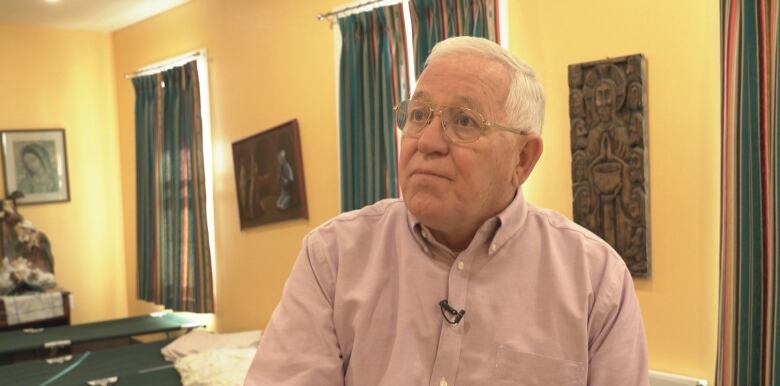 A white-haired man wearing glasses and a pink collared shirt poses for a photo, with a row of cots shown behind him.