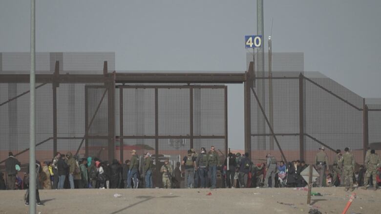 A large cluster of people line up in front of a tall metal gate, as guards with guns patrol the area.