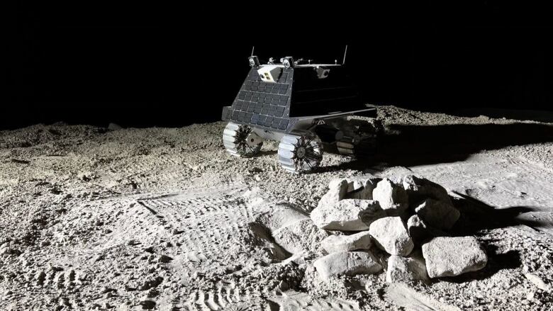 A black and grey robot with wheels sits on a rocky surface. 