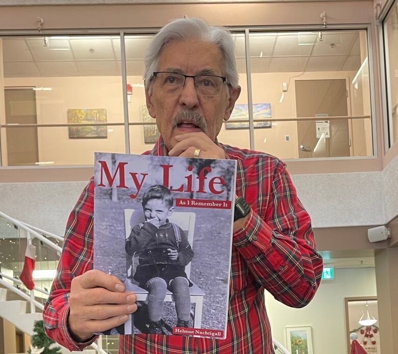 A man with silver hair, glasses and wearing a red plaid shirt holds up a book that says 