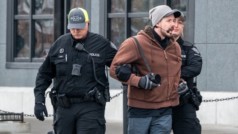 A man is flanked by two Calgary police officers, one on each arm, who appear to be marching him away from a building. 