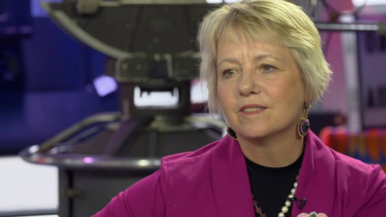 A middle-aged white woman with short blonde hair is framed from the shoulders up. She wears a fuchsia blazer, beaded brooch and earrings and a pearl necklace.