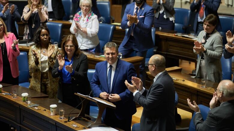 A smiling man in a blue suit stands in a legislative chamber, while other people stand around him applauding. 