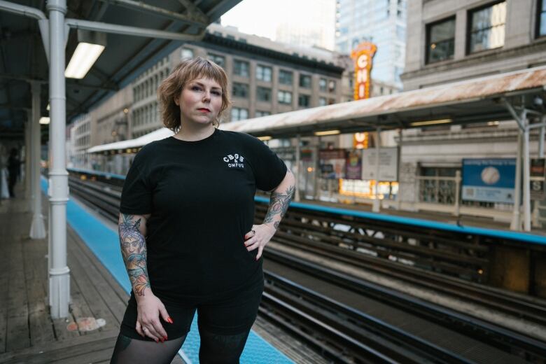 A woman with short hair wearing all black stands in front of a train track.