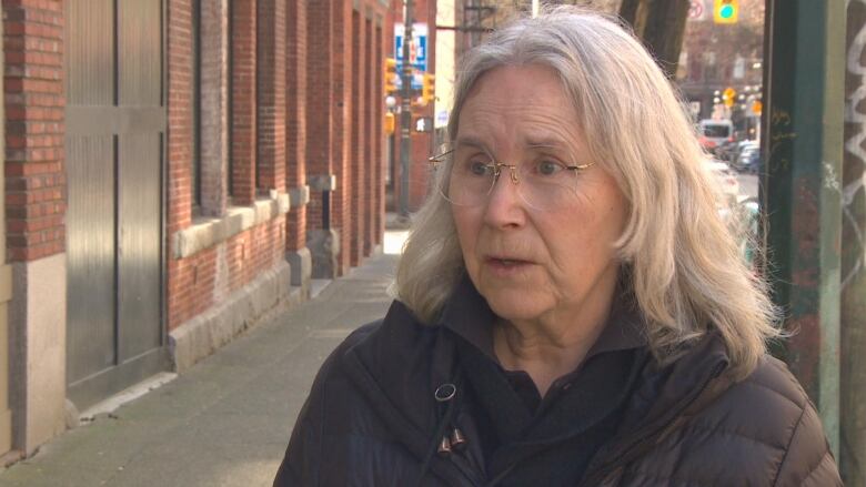 A white woman with shoulder-length white hair talks while standing on a pavement.