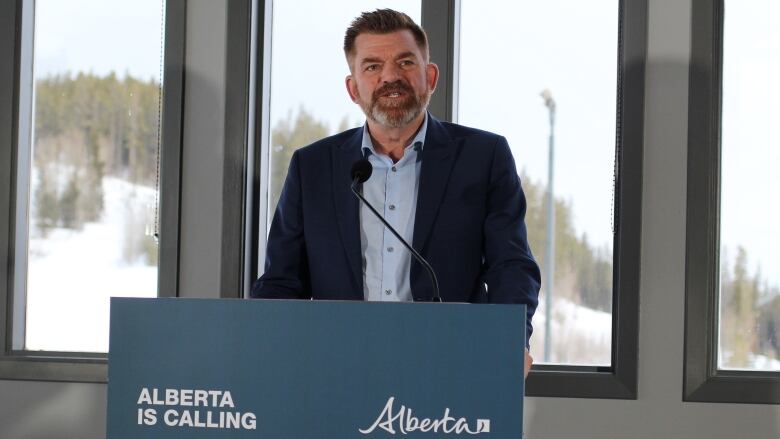 A man wearing a blue suit and light blue shirt stands and speaks at a podium that has a sign that says Alberta is Calling.