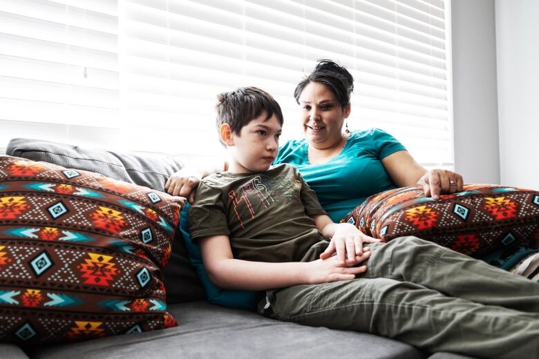 a mother sits with her son on the couch in front of a bright window.