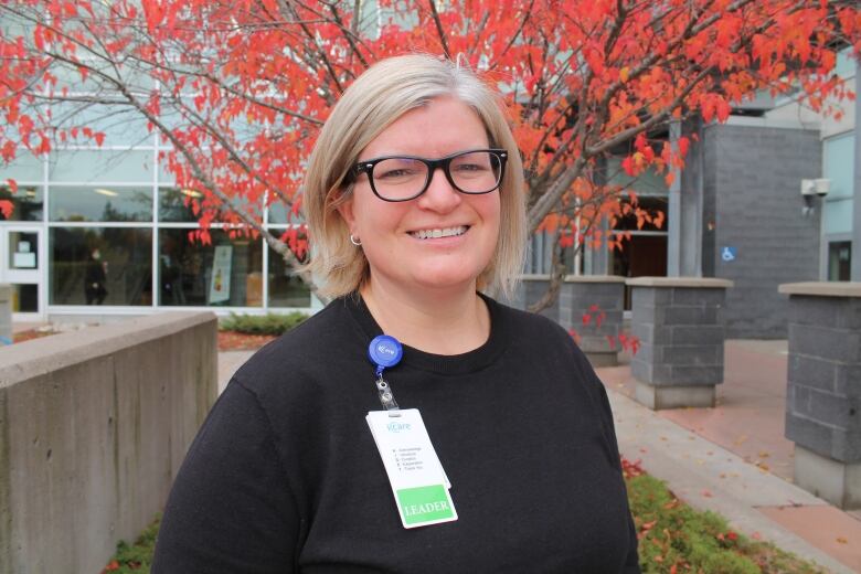 Woman with blonde hair and glasses, wearing a black shirt and name tag stands in front of a tree.