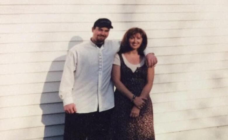 Man in white dress shirt and ball cap drapes his arm around woman in brown dress.