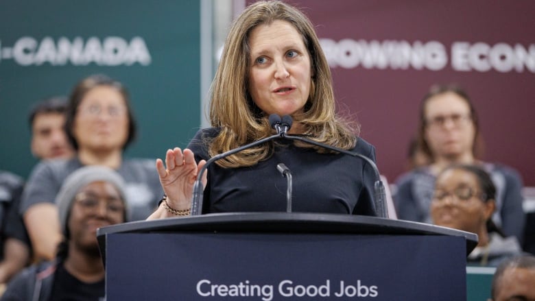 White woman with shoulder length hair speaks at a podium and gestures with her right hand.